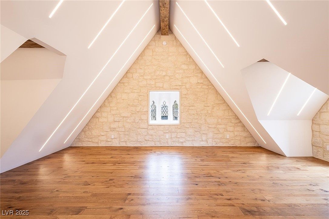 This is an interior shot of a finished attic space. The room features hardwood flooring, a stone accent wall with a window, and recessed lighting. The overall impression is a clean, modern, and well-lit bonus space.