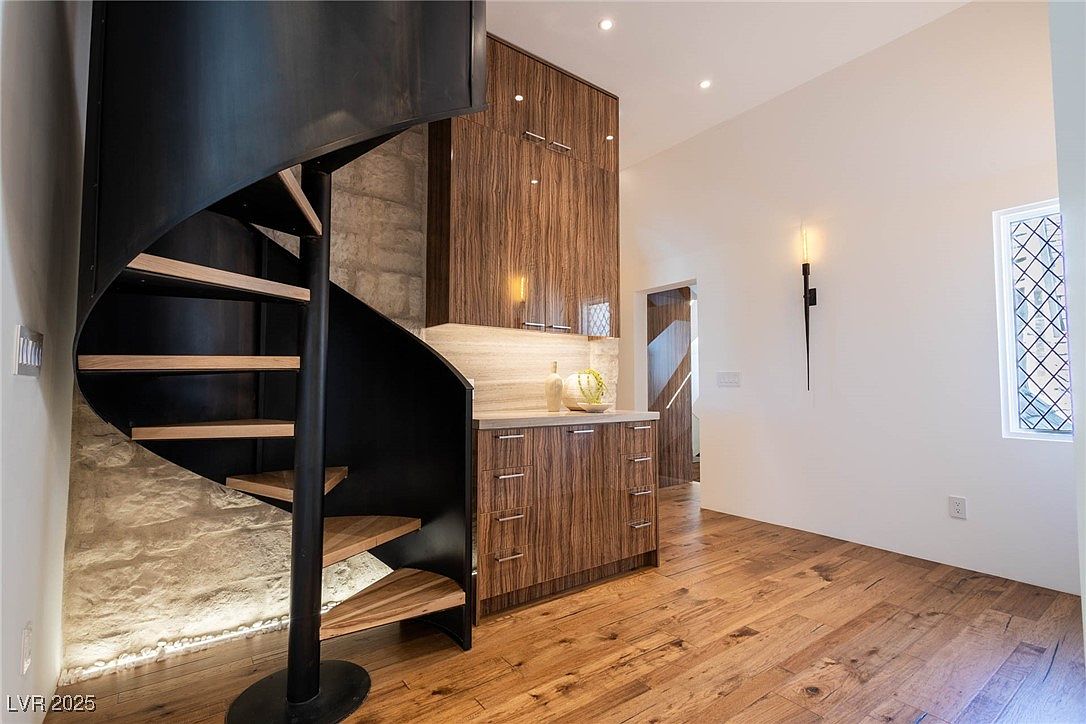 This interior shot showcases a modern hallway featuring a striking spiral staircase with wooden steps and a dark metal frame. Adjacent to the staircase is a built-in cabinet with a countertop, adding functionality to the space. The hardwood flooring and natural light from a nearby window create a warm and inviting atmosphere.