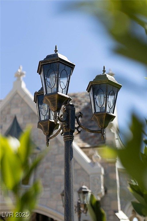 The image showcases a decorative, vintage-style lamppost with three lanterns, set against the backdrop of a stone building with architectural details. The lamppost features ornate metalwork and glass panels, adding a touch of elegance and charm. The scene is framed by foliage, creating a sense of depth and highlighting the fixture as a key element of the property's exterior design.