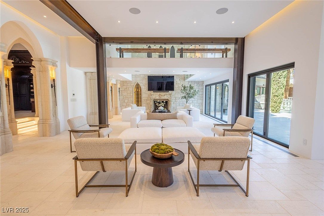 This is an interior shot of a luxurious living room featuring a neutral color palette with white walls and light tile flooring. The room is furnished with modern seating arrangements, including a white sofa and several armchairs. A striking architectural feature is the steel beam and glass partition that separates the living area from an upper level, adding a contemporary touch to the space.