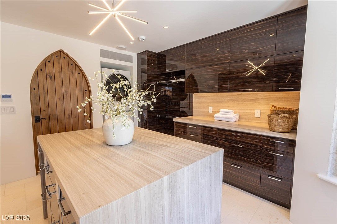 This interior shot showcases a modern laundry room with high-end finishes. Dark wood cabinetry with sleek hardware lines the walls, complemented by a light countertop and backsplash. A unique wooden arched door adds character, while a front-loading washer and dryer are discreetly integrated into the design. The room is well-lit with modern fixtures, creating a clean and sophisticated space.