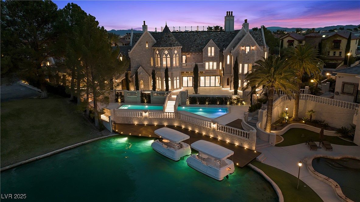 This aerial view showcases a luxurious estate at dusk, featuring a grand stone mansion with multiple pools and a private lake. Two boats are docked in the illuminated water, adding to the property's unique appeal. The manicured landscaping, architectural details, and overall ambiance suggest high-end living and exclusivity.