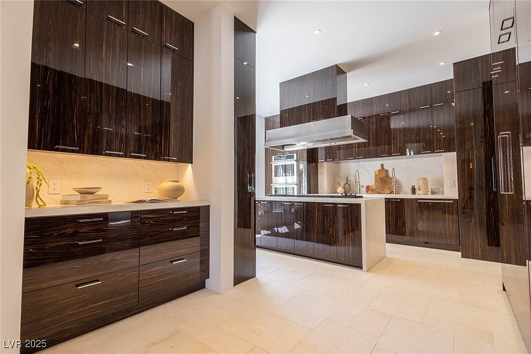 This is a modern kitchen with dark wood cabinetry and light countertops. The kitchen features a central island and stainless steel appliances. The overall impression is sleek and luxurious, with a focus on clean lines and high-end finishes.