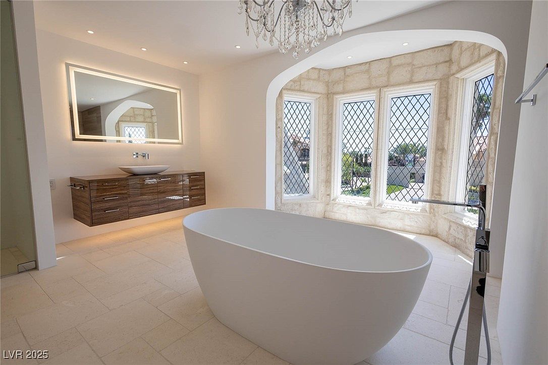 This is a luxurious primary bathroom featuring a freestanding white bathtub as the focal point. A floating vanity with a vessel sink and a large, illuminated mirror add to the modern aesthetic. The room is brightened by a bay window with diamond-patterned panes, and a chandelier hangs from the ceiling, enhancing the elegant ambiance.