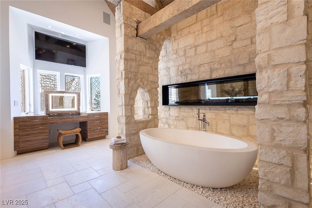 This is a luxurious primary bathroom featuring a freestanding oval bathtub set on a bed of pebbles, framed by a stone archway with a built-in fireplace. A vanity with a lighted mirror and a television above it are visible in the background, complemented by decorative windows. The room exudes a spa-like ambiance with its neutral color palette and high-end finishes.
