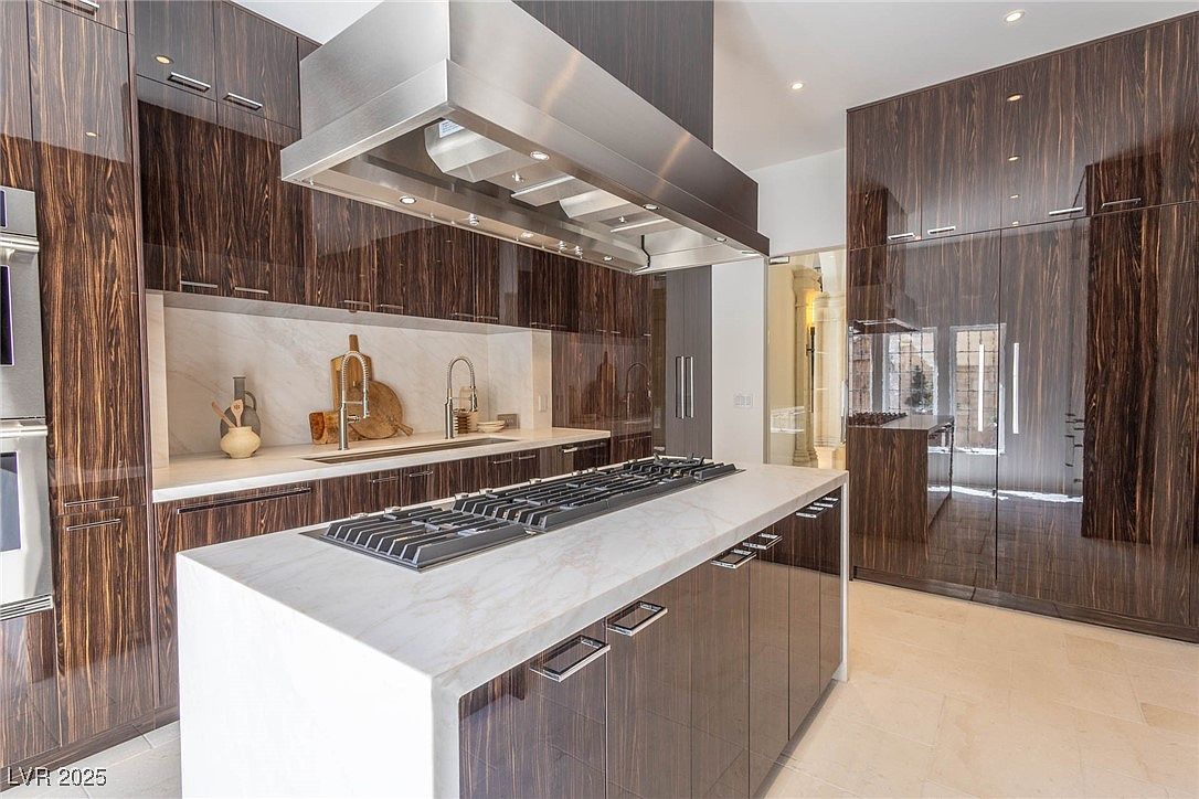 This is a modern kitchen with dark wood cabinetry and a large island featuring a cooktop and white countertops. Stainless steel appliances and a large range hood add to the contemporary feel. The kitchen has a sleek and luxurious design, with a focus on clean lines and high-end finishes.