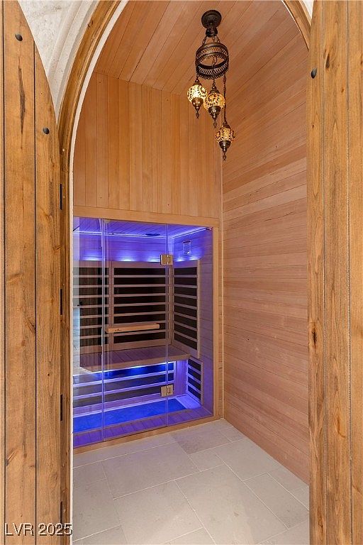 This image showcases a modern sauna room, featuring light wood paneling on the walls and ceiling, creating a warm and inviting atmosphere. The sauna itself is enclosed in glass, with blue lighting adding a contemporary touch. An ornate chandelier hangs from the ceiling, providing a unique focal point.