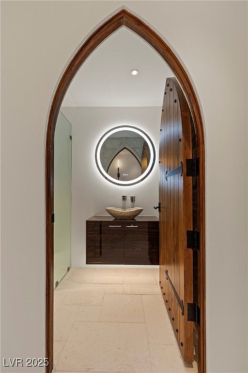 This image showcases a unique bathroom design, viewed through a gothic archway. The bathroom features a modern, round, backlit mirror above a floating vanity with a vessel sink. A partially open wooden door with iron hardware adds a rustic touch, contrasting with the contemporary fixtures and light-colored tile flooring.