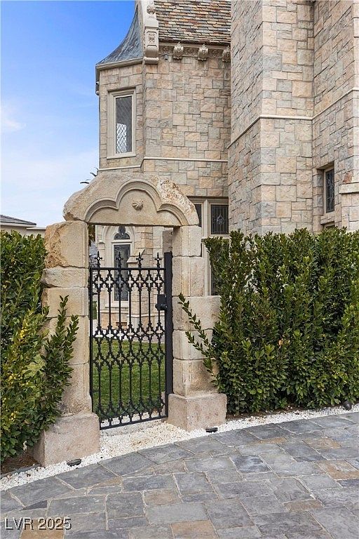 This image showcases an elegant entryway featuring a stone archway and a wrought iron gate, leading into a lush green space. The exterior of the building is constructed with light-colored stone, suggesting a stately and sophisticated design. The pathway is paved with large stone tiles, complemented by white gravel accents, enhancing the overall curb appeal.