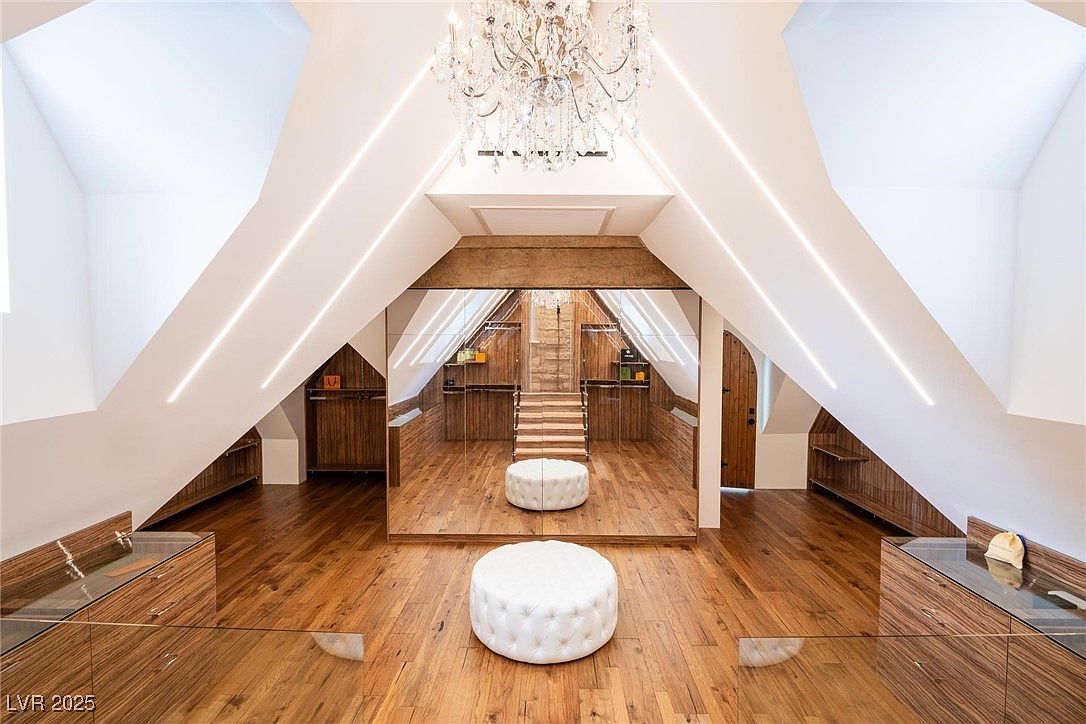 This is a luxurious walk-in closet featuring hardwood floors, custom wood cabinetry, and a central mirrored wall that creates an illusion of depth. A crystal chandelier hangs from the ceiling, adding a touch of elegance, while white ottomans provide seating. The space is well-lit with modern, linear lighting fixtures.