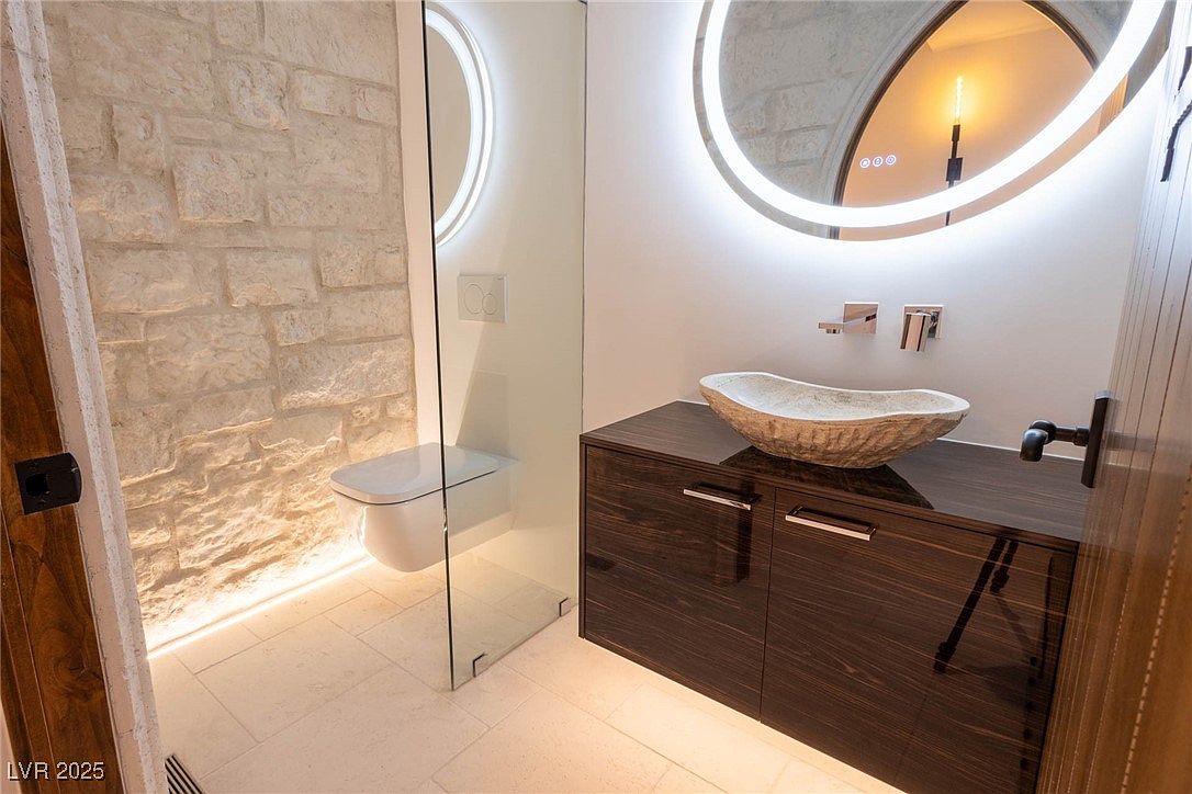 This is a modern bathroom featuring a stone accent wall, a floating toilet, and a vessel sink on a dark wood vanity. A circular, illuminated mirror adds a touch of elegance, and the glass shower enclosure enhances the room's spacious feel. The overall design is sleek and contemporary.