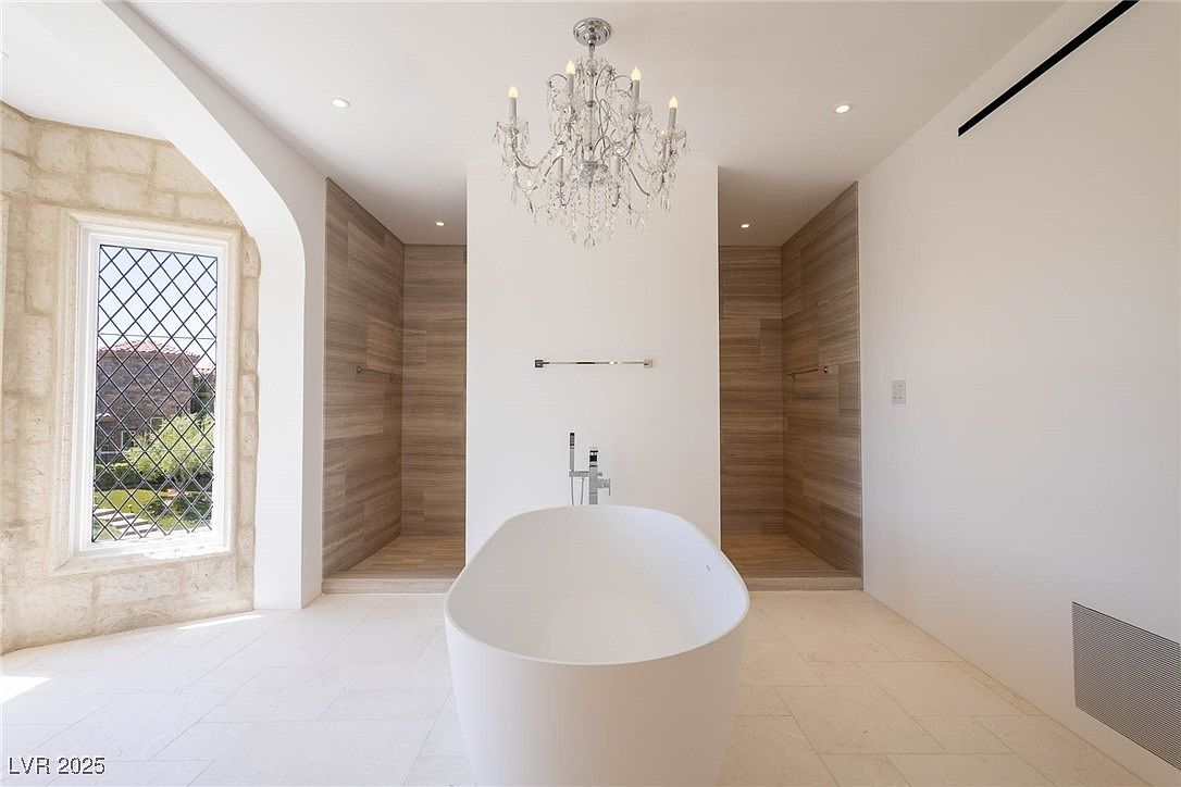 This is a luxurious primary bathroom featuring a freestanding white bathtub as the focal point, positioned between two walk-in showers with wood-look tiling. A crystal chandelier hangs above the tub, adding a touch of elegance. The room is bright and spacious, with a large window providing natural light and a view to the outside.