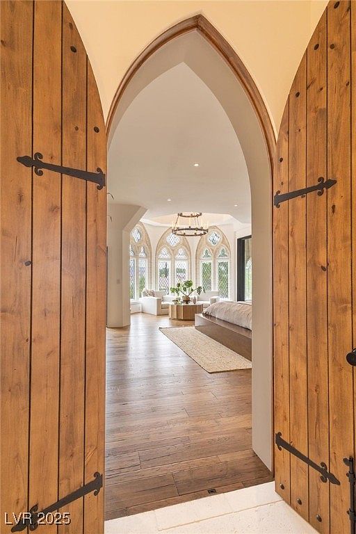 This image showcases a luxurious primary bedroom viewed through an arched doorway with rustic wooden doors. The room features a unique architectural design with arched windows, a chandelier, and a neutral color palette. The hardwood flooring and area rug add warmth and texture to the space, creating an inviting and elegant atmosphere.