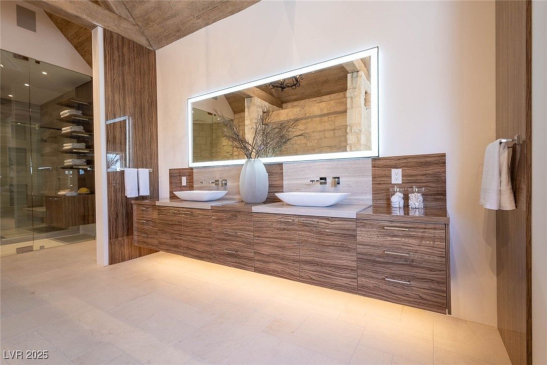 This is a primary bathroom featuring a double vanity with vessel sinks and modern faucets. The vanity has wood-grain cabinets and drawers, and a large, illuminated mirror hangs above. The bathroom has a contemporary design with clean lines and neutral colors, and a glass-enclosed shower is visible to the left.