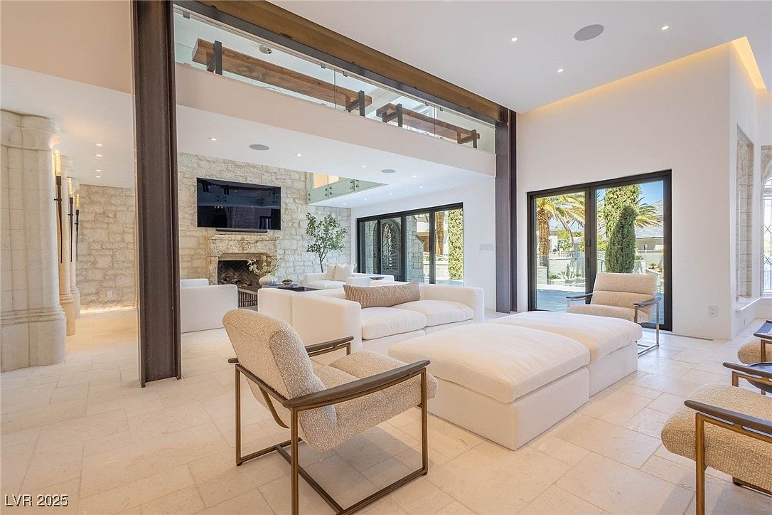 This is a spacious living room featuring a stone accent wall with a fireplace and a large television. The room is furnished with white sofas, ottomans, and modern chairs with metal frames. Large sliding glass doors provide ample natural light and access to the exterior, creating an open and airy atmosphere.