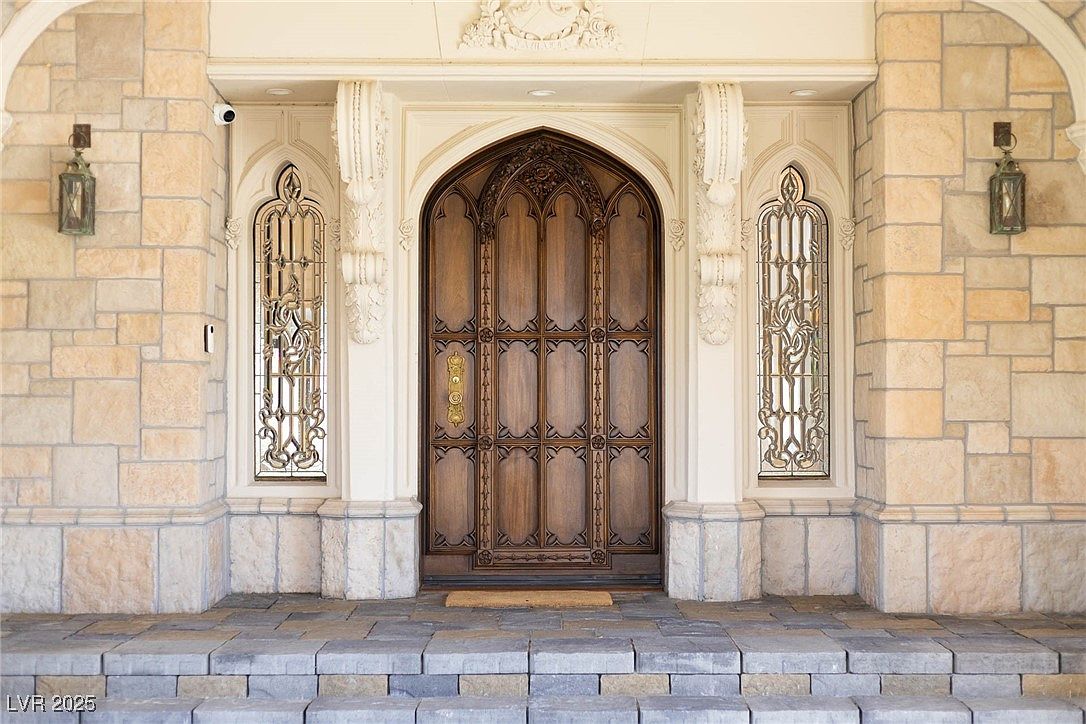 This image showcases a grand entryway with a large, ornate wooden door as the focal point. The entrance is framed by decorative stone pillars and arched windows, adding to the architectural detail. The steps leading up to the door are made of stone, creating a stately and inviting entrance.