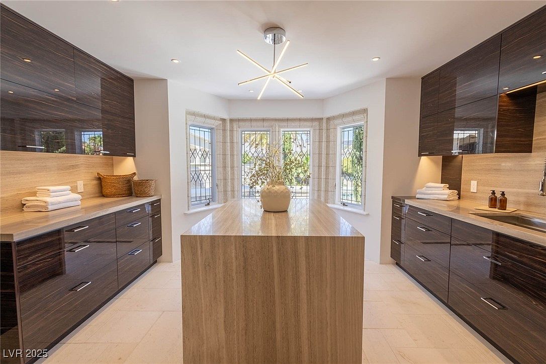 This is a well-lit kitchen featuring dark wood cabinetry and light-colored countertops. A central island with a matching countertop is adorned with a decorative vase. The kitchen is illuminated by a modern chandelier and natural light streaming through a bay window, creating a bright and inviting atmosphere.