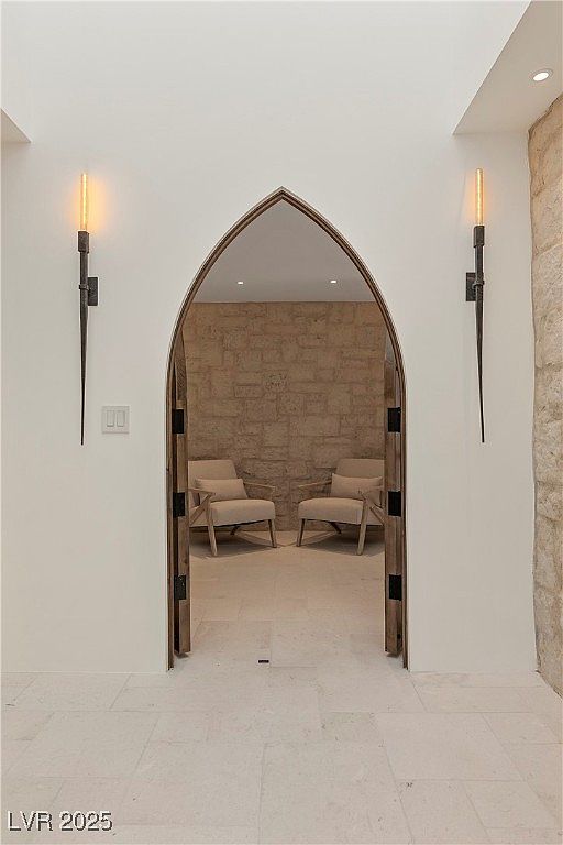 This interior shot showcases a unique arched doorway leading to a cozy sitting area. The hallway features light-colored stone flooring and sconce lighting, while the sitting area has a textured stone wall and two armchairs. The design creates an inviting and sophisticated atmosphere.