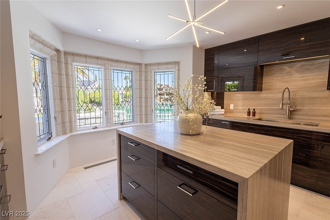 This is a well-lit kitchen featuring dark wood cabinetry and a light-colored countertop island. The kitchen has a modern design with a unique lighting fixture and a large window offering natural light. The overall impression is clean and sophisticated, highlighting the kitchen's functionality and style.