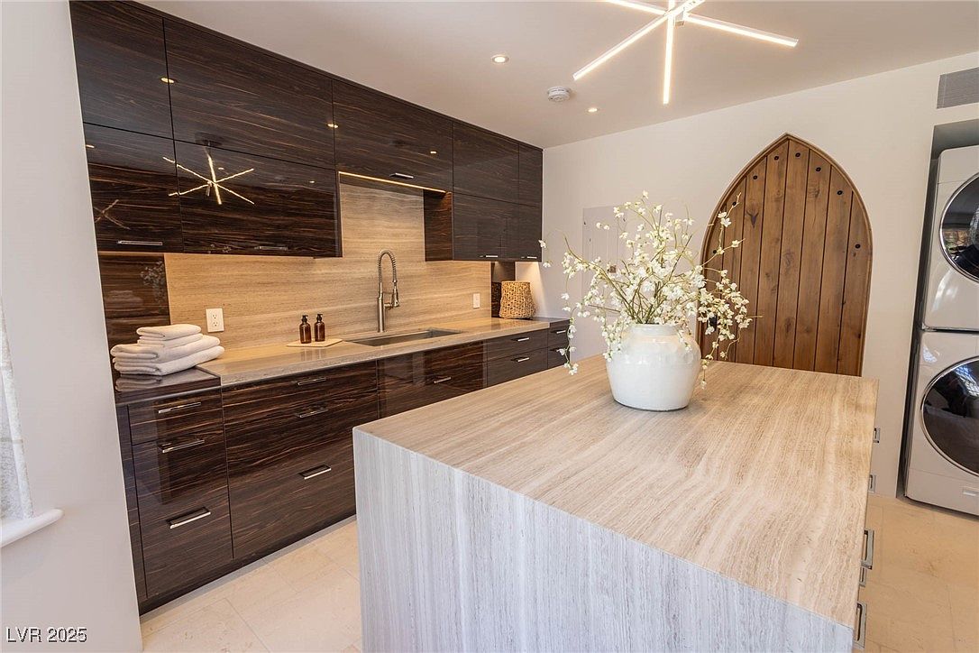 This interior shot showcases a modern laundry room with dark wood cabinetry and a light-colored countertop. A central island with a light wood finish provides ample workspace, while a unique arched wooden door adds a touch of character. Stacked washer and dryer units are visible on the right, and the overall impression is one of sophisticated functionality.