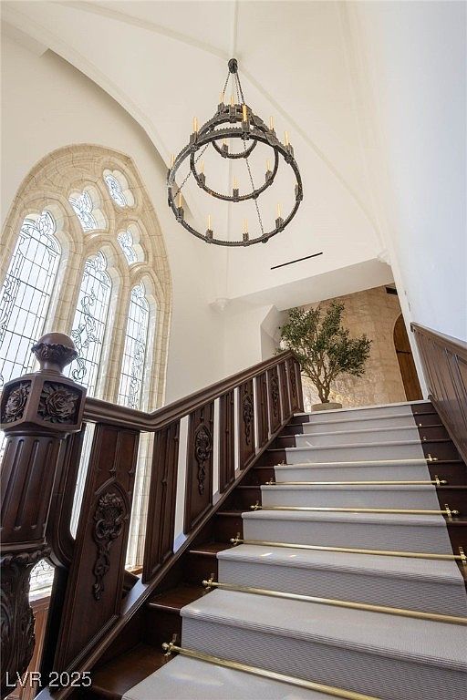 This interior shot showcases a grand staircase with dark wood railings and white carpeted steps accented with gold rods. A large arched window provides natural light, complemented by an ornate chandelier hanging from the high ceiling. The overall impression is one of elegance and architectural detail.