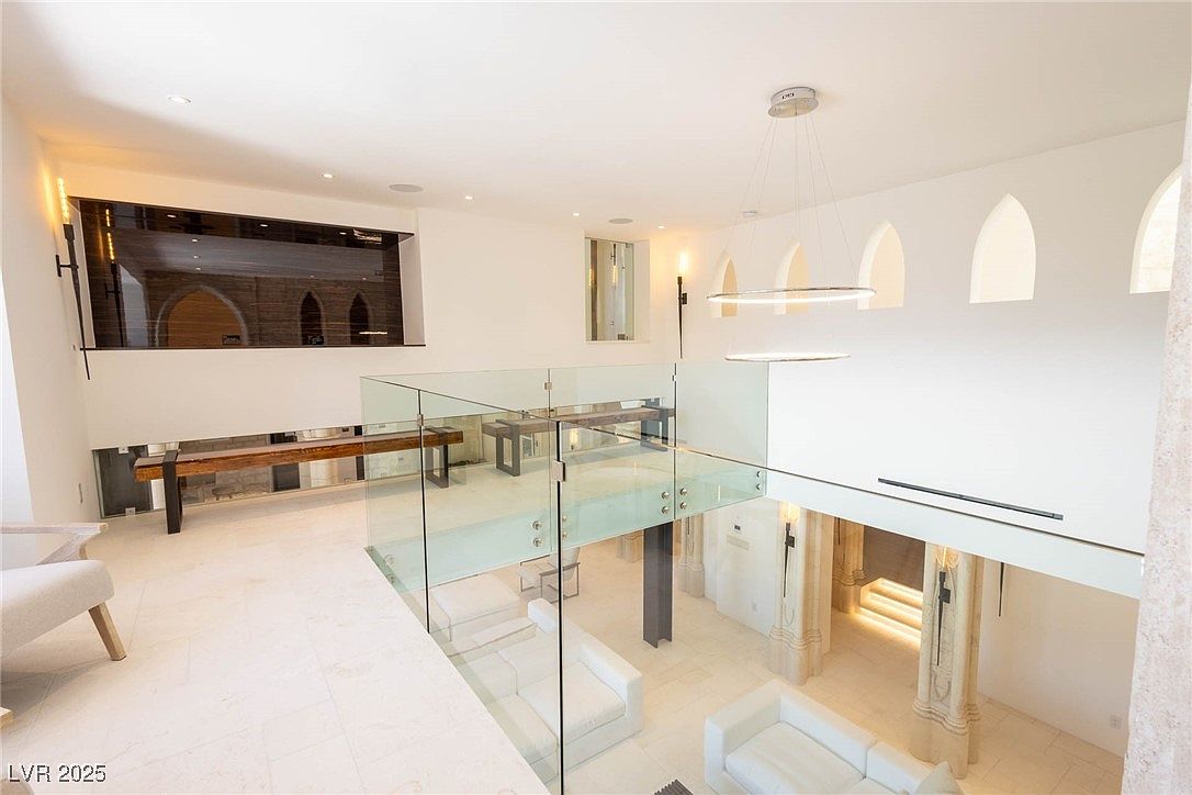 This interior shot showcases a modern hallway and staircase area with a clean, minimalist aesthetic. The space features white walls, light-colored flooring, and glass railings, creating an open and airy feel. A unique architectural window and modern lighting fixtures add visual interest, enhancing the contemporary design.