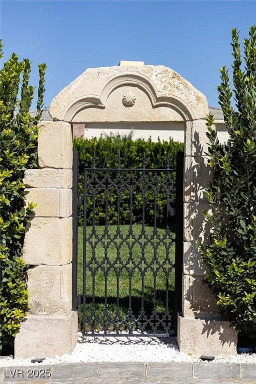 The image showcases an elegant entryway featuring a stone archway and a decorative wrought iron gate. Lush greenery flanks the entrance, adding a touch of natural beauty. The overall impression is one of sophistication and privacy, suggesting a well-maintained and upscale property.