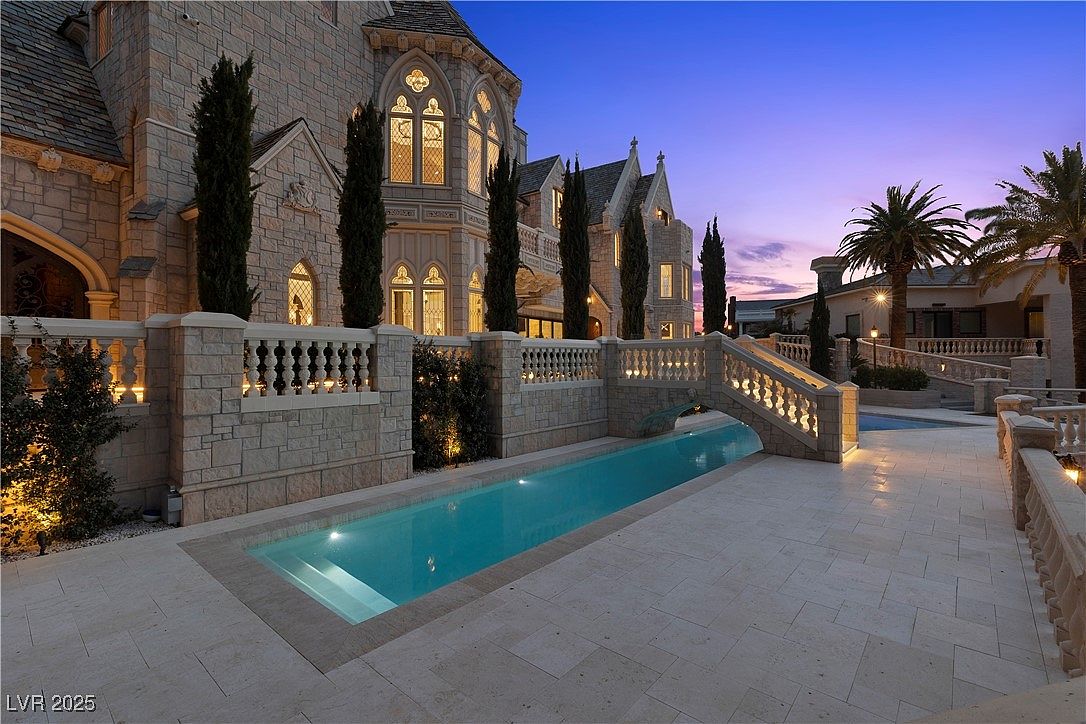 This exterior shot showcases a luxurious pool area at dusk, featuring a long, narrow pool with a bridge over it, connecting to a grand stone building with arched windows and manicured landscaping. The scene is illuminated by subtle lighting, highlighting the architectural details and creating an elegant and inviting atmosphere, perfect for a high-end real estate listing.