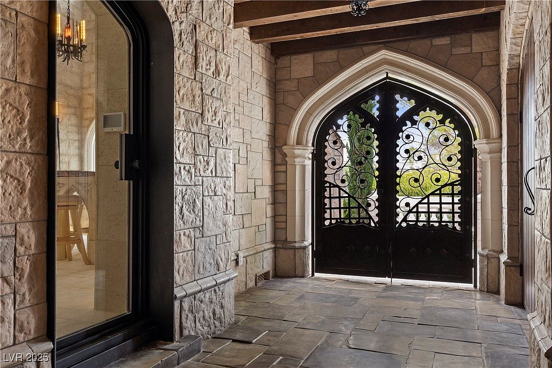 This image showcases a grand entryway featuring a stone-paved walkway leading to an ornate, wrought-iron gate set within a gothic-style archway. The walls are constructed of textured stone, adding to the castle-like aesthetic. To the left, a glass door offers a glimpse into the interior, where a chandelier is visible, enhancing the luxurious feel of the property.