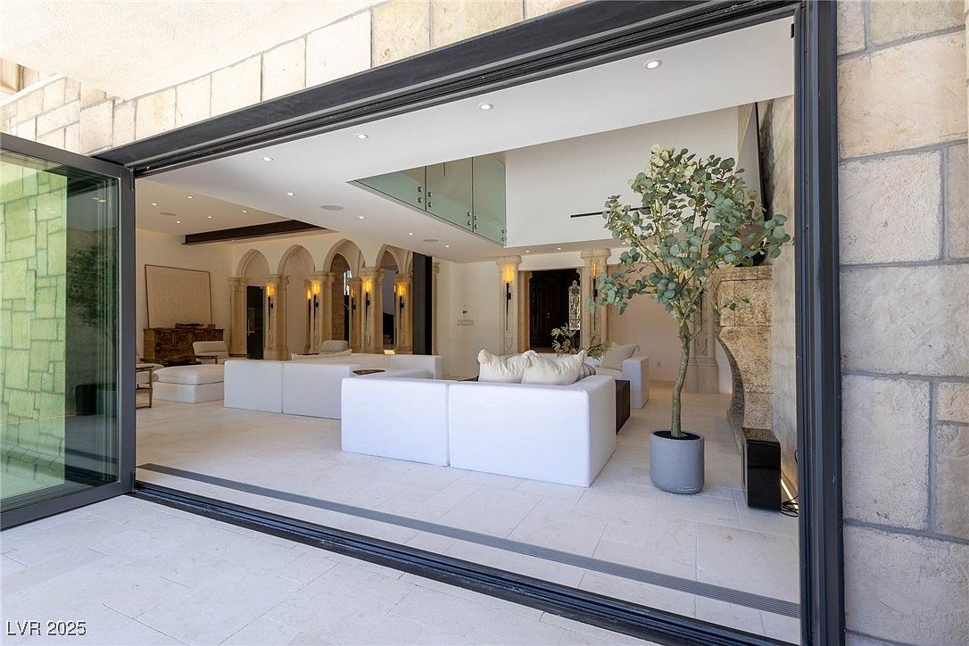 This image showcases a luxurious living room with a seamless indoor-outdoor transition through large, open sliding glass doors. The interior features a modern design with white sofas, stone columns, and elegant lighting fixtures. The open floor plan and neutral color palette create a spacious and inviting atmosphere, perfect for entertaining or relaxing.