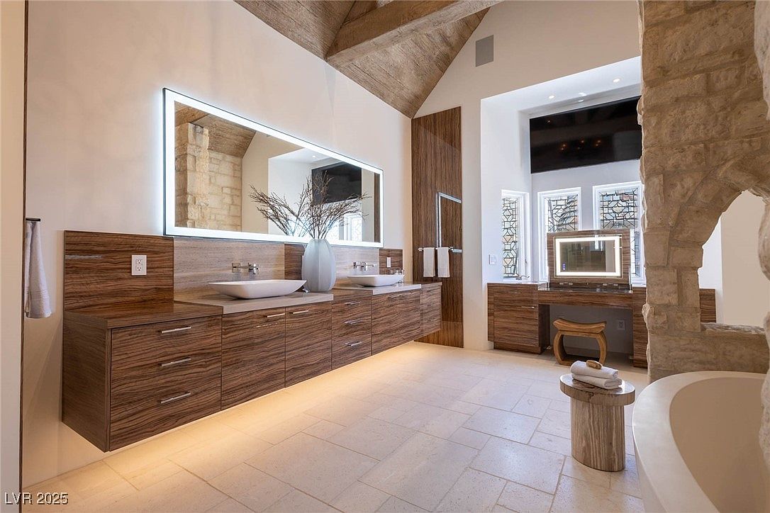 This is a luxurious primary bathroom featuring a double vanity with modern sinks and a large, illuminated mirror. The cabinetry is a rich wood grain, and the flooring is light-colored tile. A separate vanity area with a lighted mirror and stool is visible, along with a stone archway leading to the bathtub area, creating a spa-like atmosphere.