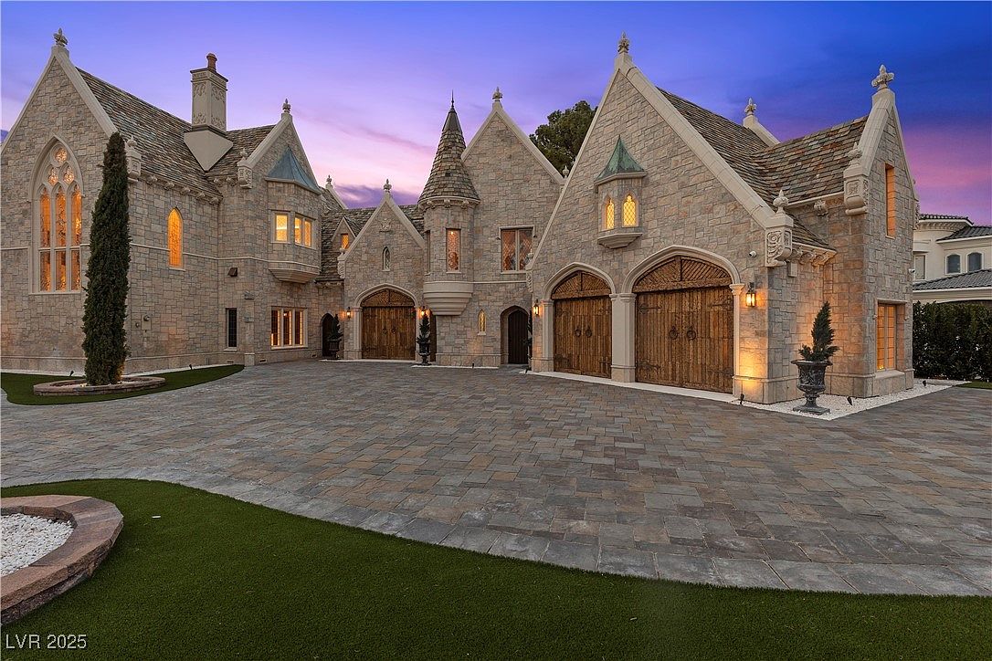This is a front exterior view of a grand, castle-like home. The architecture features stone construction, arched garage doors, and multiple gabled roofs. The driveway is paved with stone, and there is a manicured lawn in the foreground, creating an impressive and luxurious curb appeal.