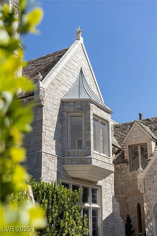 The image showcases the front exterior of a stately stone house with distinct architectural details. A prominent bay window with leaded glass adds character, while the steeply pitched roof and decorative finials contribute to the home's elegant aesthetic. The well-maintained facade and lush greenery suggest a property of considerable value and curb appeal.