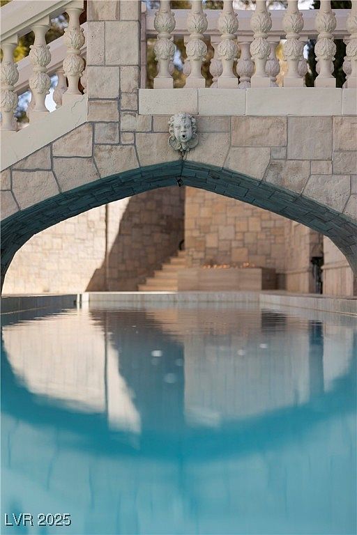 This image showcases a luxurious pool area, featuring a stone bridge with an ornate balustrade and a decorative lion head fountain. The clear blue water reflects the architectural details, creating a serene and elegant atmosphere. The stone work and design elements suggest a high-end property with a focus on aesthetic appeal.