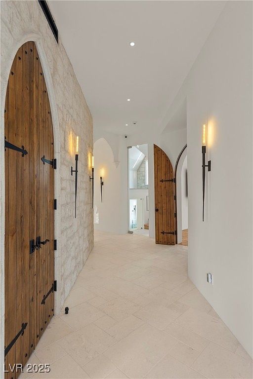 This interior shot showcases a unique hallway featuring arched doorways and custom wooden doors with iron hardware, creating a medieval-inspired aesthetic. The walls are a mix of textured stone and smooth white surfaces, illuminated by sconce lighting that mimics torches. The flooring is light-colored tile, and the perspective is a straight-on view down the hallway.