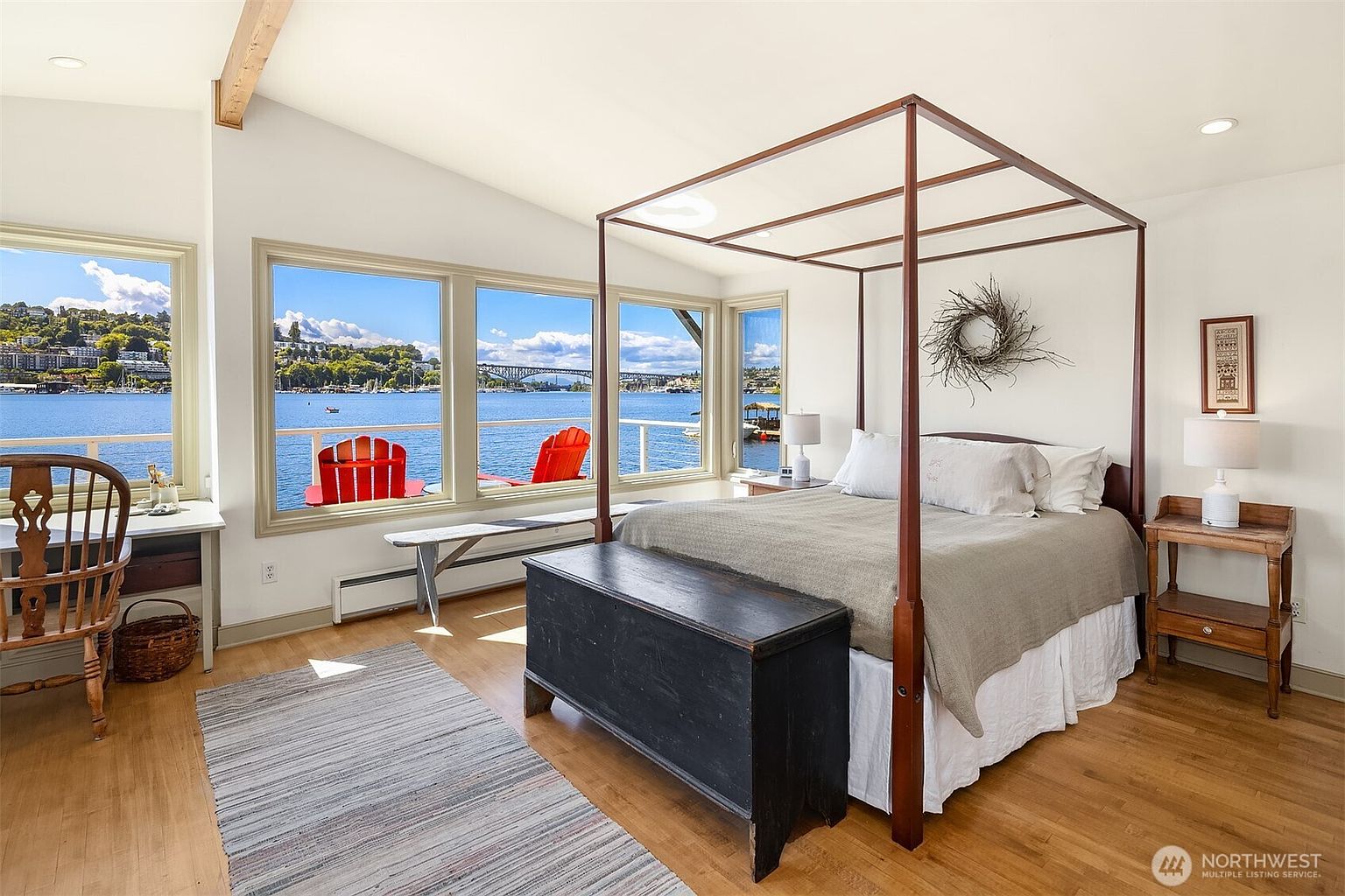This is a bright and airy primary bedroom featuring a four-poster bed with neutral bedding, a dark wood chest at the foot of the bed, and a view of the water through large windows. The room has hardwood floors, a light-colored rug, and a desk area, creating a serene and inviting atmosphere. The style is a blend of traditional and contemporary, emphasizing comfort and natural light.