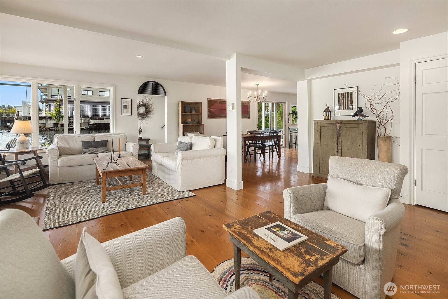 This inviting living room features hardwood floors, comfortable white sofas, and rustic wooden coffee tables, creating a cozy and relaxed atmosphere. The open layout connects to the dining area, enhancing the sense of spaciousness, while natural light streams in through the windows, offering a glimpse of the waterfront. A mix of textures and neutral tones contributes to the room's warm and welcoming appeal.
