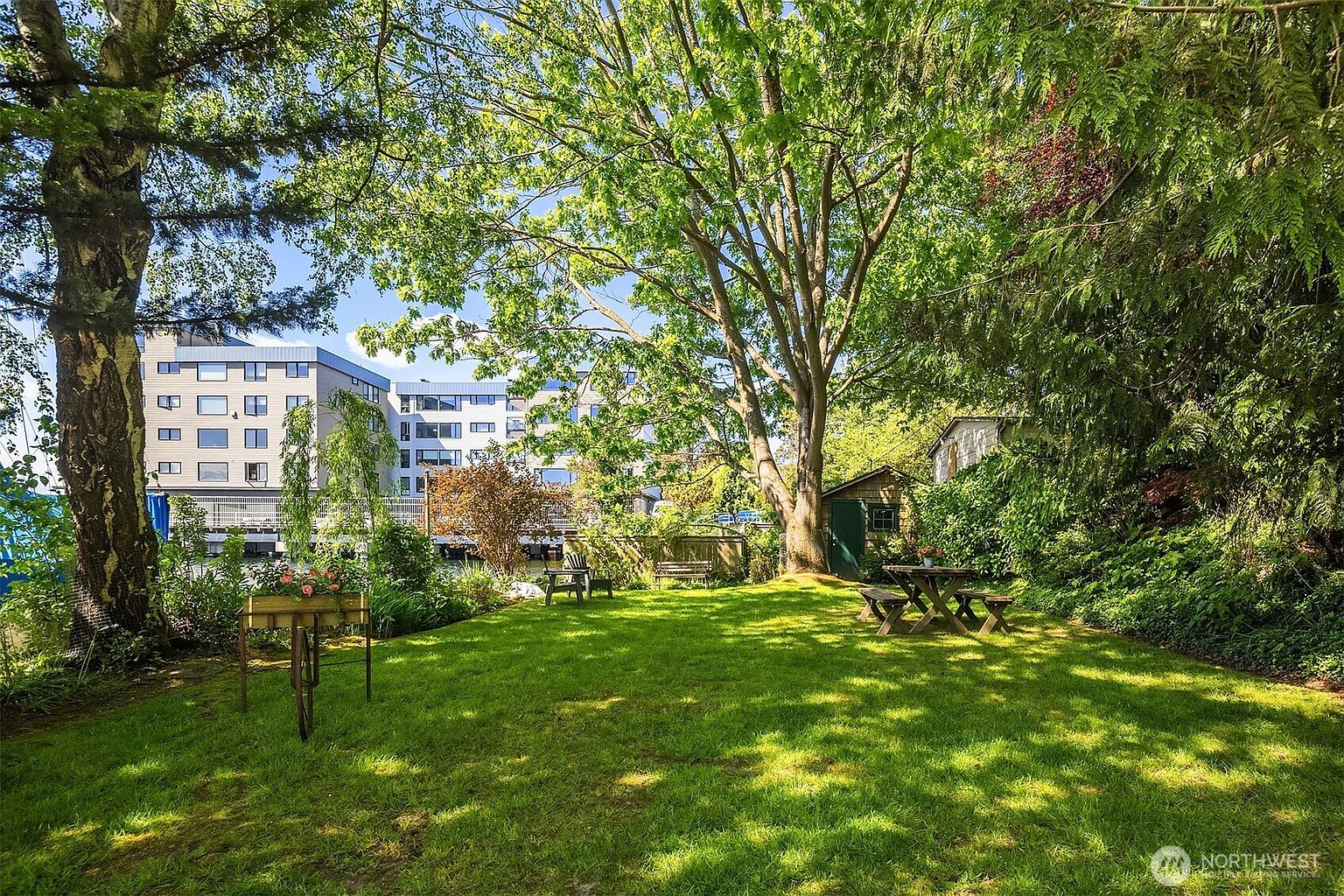 This image showcases a lush, well-maintained backyard garden. A large tree provides ample shade, and a picnic table and benches offer seating for outdoor relaxation. The garden features a mix of green lawn, mature trees, and various plants, creating a serene and inviting atmosphere.