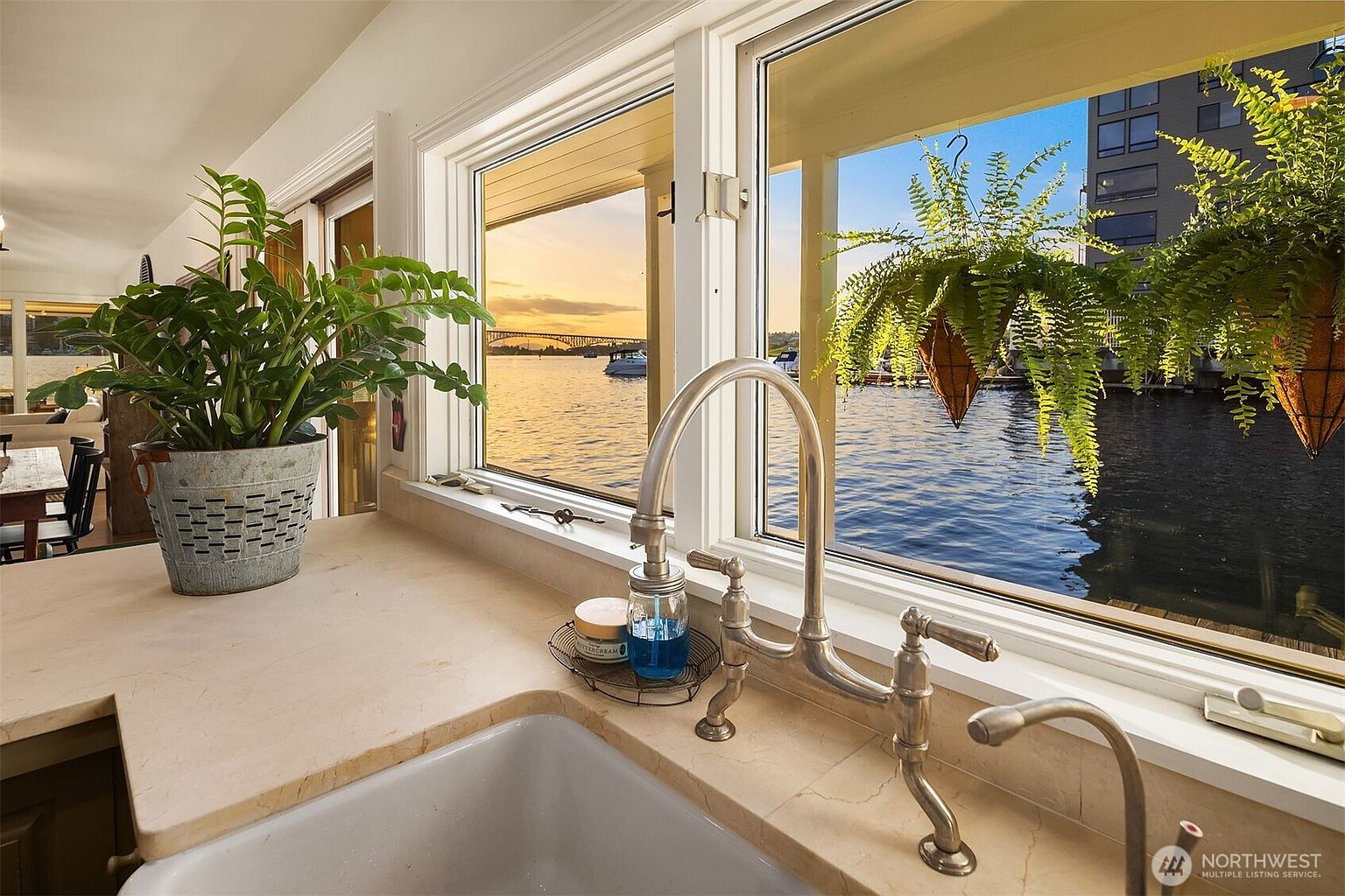 This image showcases a kitchen sink area with a large window providing a scenic view of the water and a bridge at sunset. The kitchen features a farmhouse-style sink, a vintage-looking faucet, and potted plants adding a touch of nature. The countertop is a light-colored stone, and the overall impression is one of tranquility and connection to the outdoors.