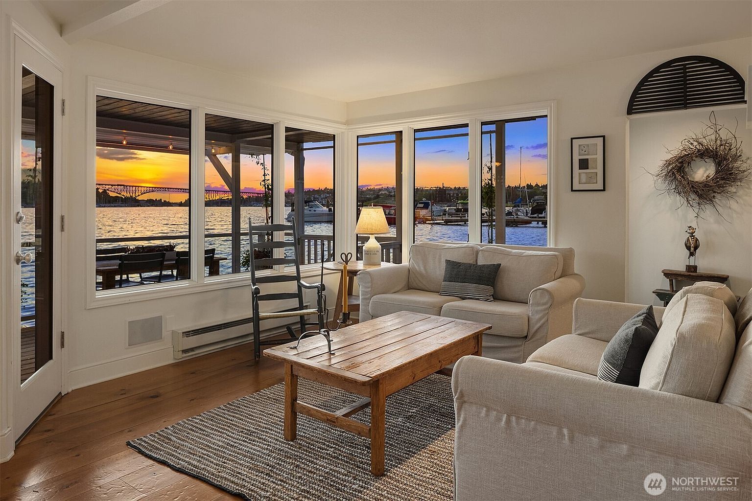 This is an inviting living room with a stunning waterfront view visible through large windows. The room features comfortable, neutral-toned sofas, a rustic wooden coffee table, and a textured area rug. The decor is tasteful and understated, creating a relaxing and elegant atmosphere, perfect for enjoying the scenic surroundings.