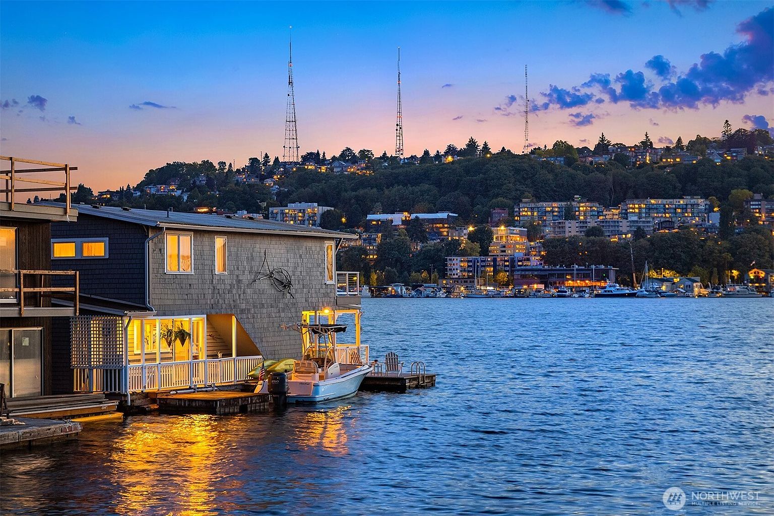 This image showcases a charming houseboat at dusk, featuring warm interior lighting that reflects beautifully on the calm water. The house is docked with a small boat, and the backdrop includes a cityscape silhouette with illuminated buildings and towers against a colorful sky. The overall impression is serene and inviting, highlighting the unique waterfront living experience.