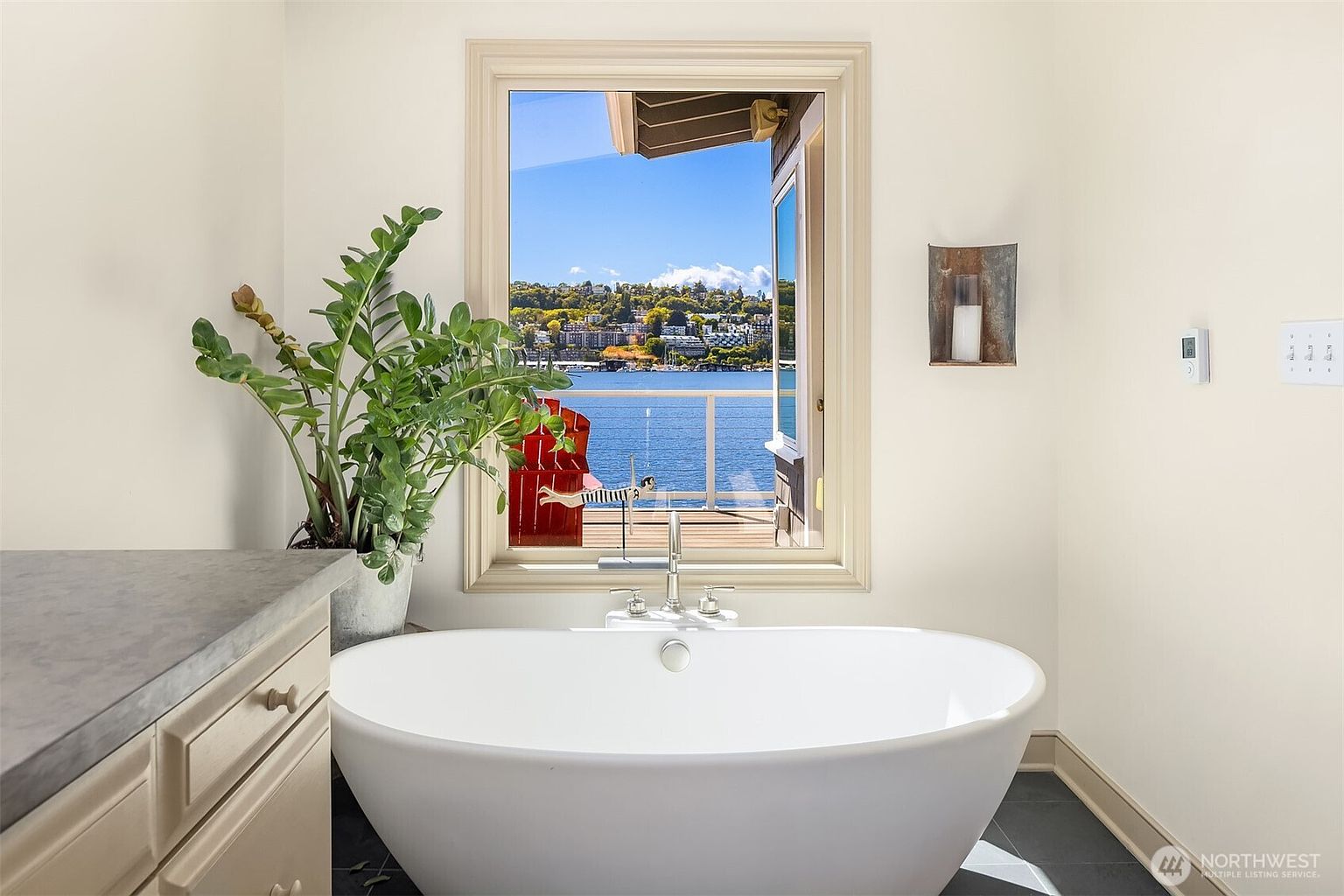 This is a bright and airy primary bathroom featuring a freestanding white bathtub positioned in front of a large window with a view of the water and cityscape. A potted plant sits on a vanity to the left of the tub, adding a touch of nature to the space. The room is painted in a neutral tone, creating a serene and spa-like atmosphere.