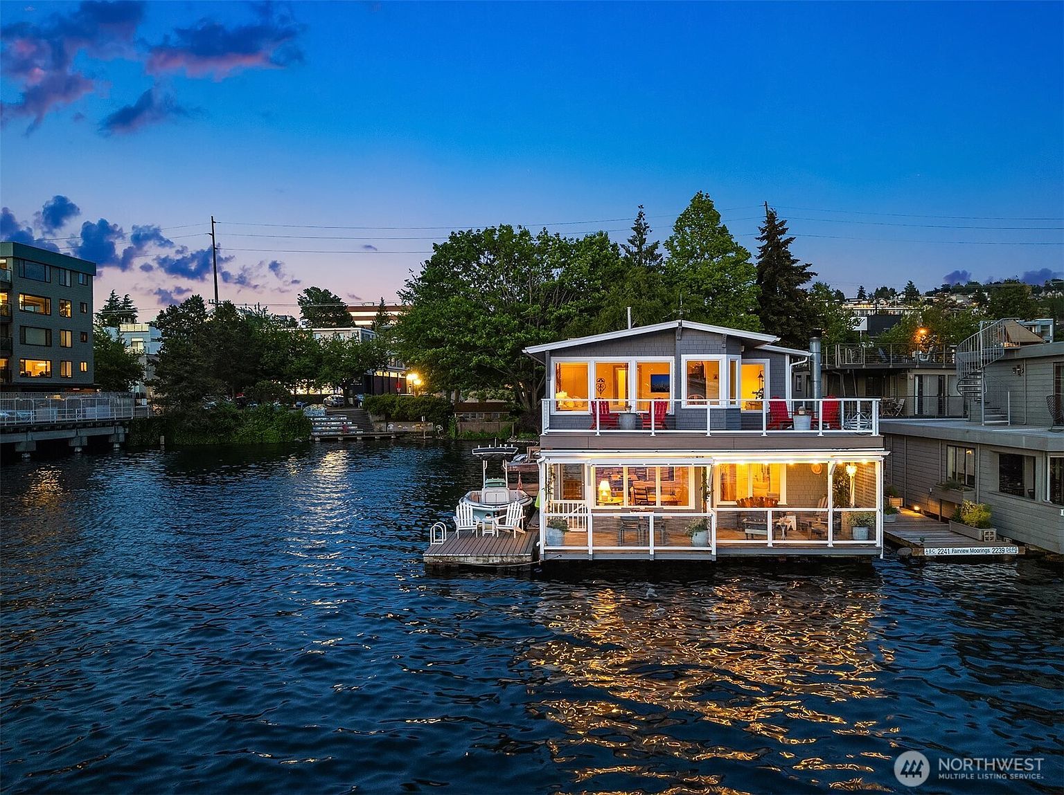 This aerial view showcases a charming two-story houseboat at dusk, featuring illuminated interiors and a cozy deck. The house is surrounded by calm water reflecting the warm lights, creating a serene and inviting atmosphere. The surrounding landscape includes trees and other waterfront properties, adding to the unique appeal of this floating home.