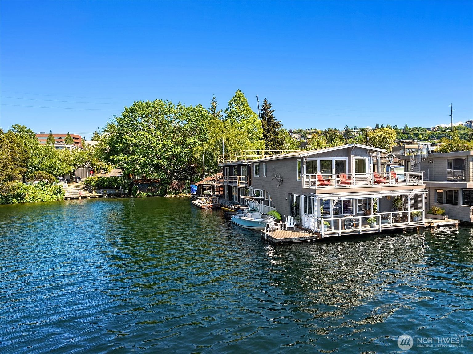 This aerial view showcases a modern houseboat with a gray exterior, multiple decks, and white railings, situated on a calm lake. The house features large windows and outdoor seating areas, creating an inviting waterfront living space. Lush greenery surrounds the property, enhancing its serene and private setting.
