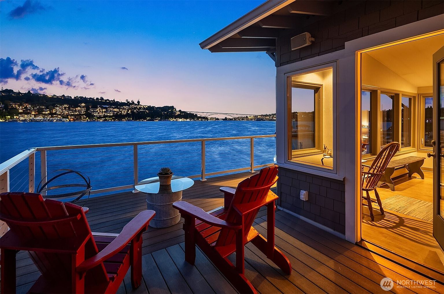 This image showcases a beautifully designed outdoor deck area with stunning waterfront views. The deck features comfortable red Adirondack chairs, a glass-topped table, and a modern railing system. An open doorway leads into a well-lit interior space, creating a seamless transition between indoor and outdoor living, perfect for enjoying the scenic surroundings.