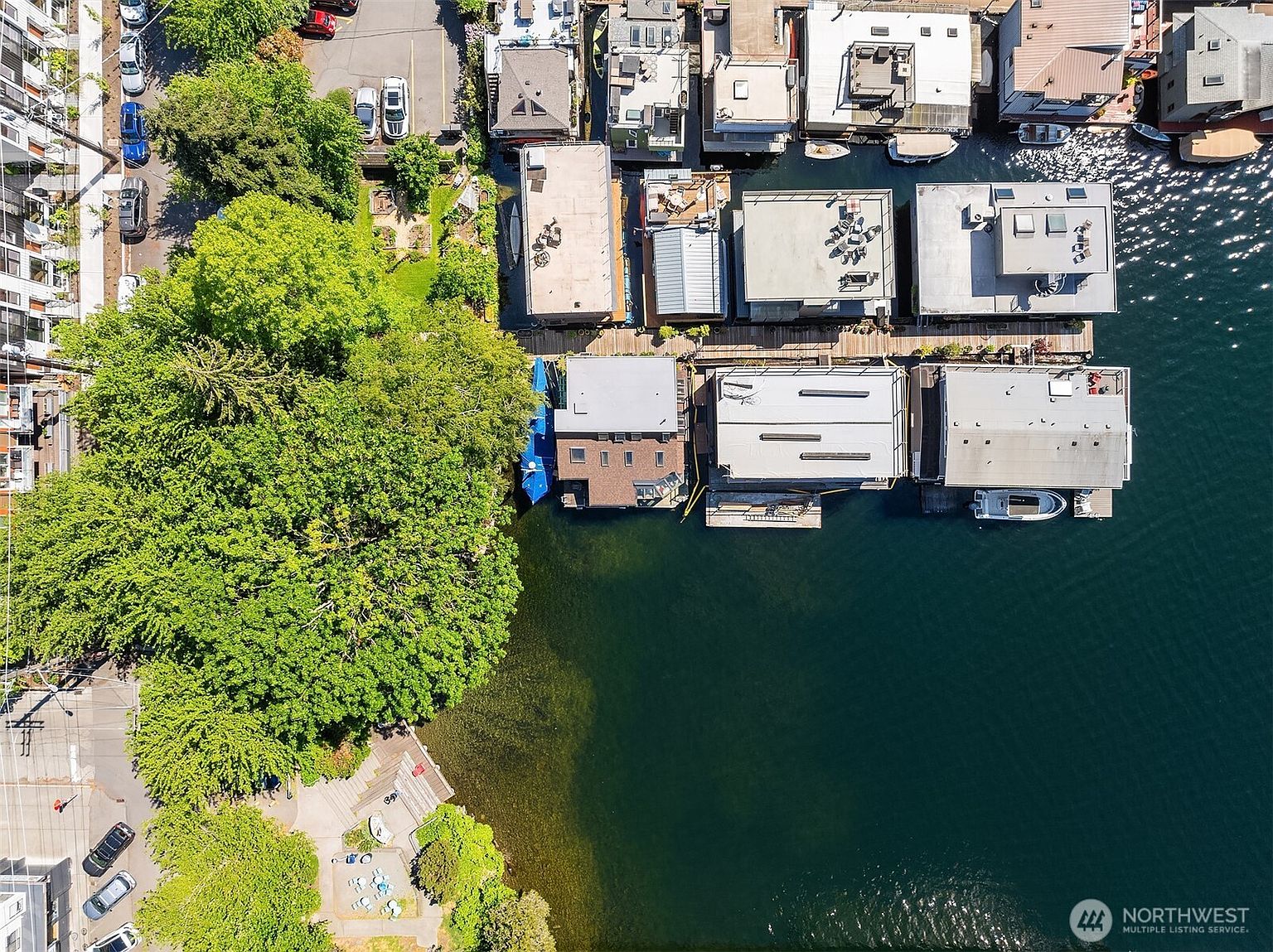 This aerial shot showcases a unique waterfront property featuring houseboats docked along a serene body of water. The rooftops of the houseboats are visible, along with boats and docks. Lush greenery lines the shore, providing a natural contrast to the urban setting and the calm water.