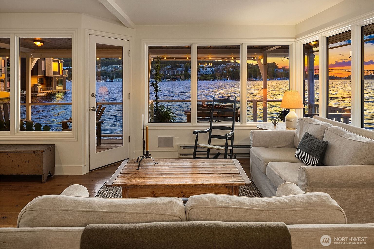 This inviting living room features a stunning waterfront view through large windows, creating a serene and relaxing atmosphere. The room is furnished with comfortable, neutral-toned sofas and a rustic wooden coffee table, complemented by a rocking chair and a table lamp. The overall style is cozy and elegant, perfect for enjoying the scenic surroundings.