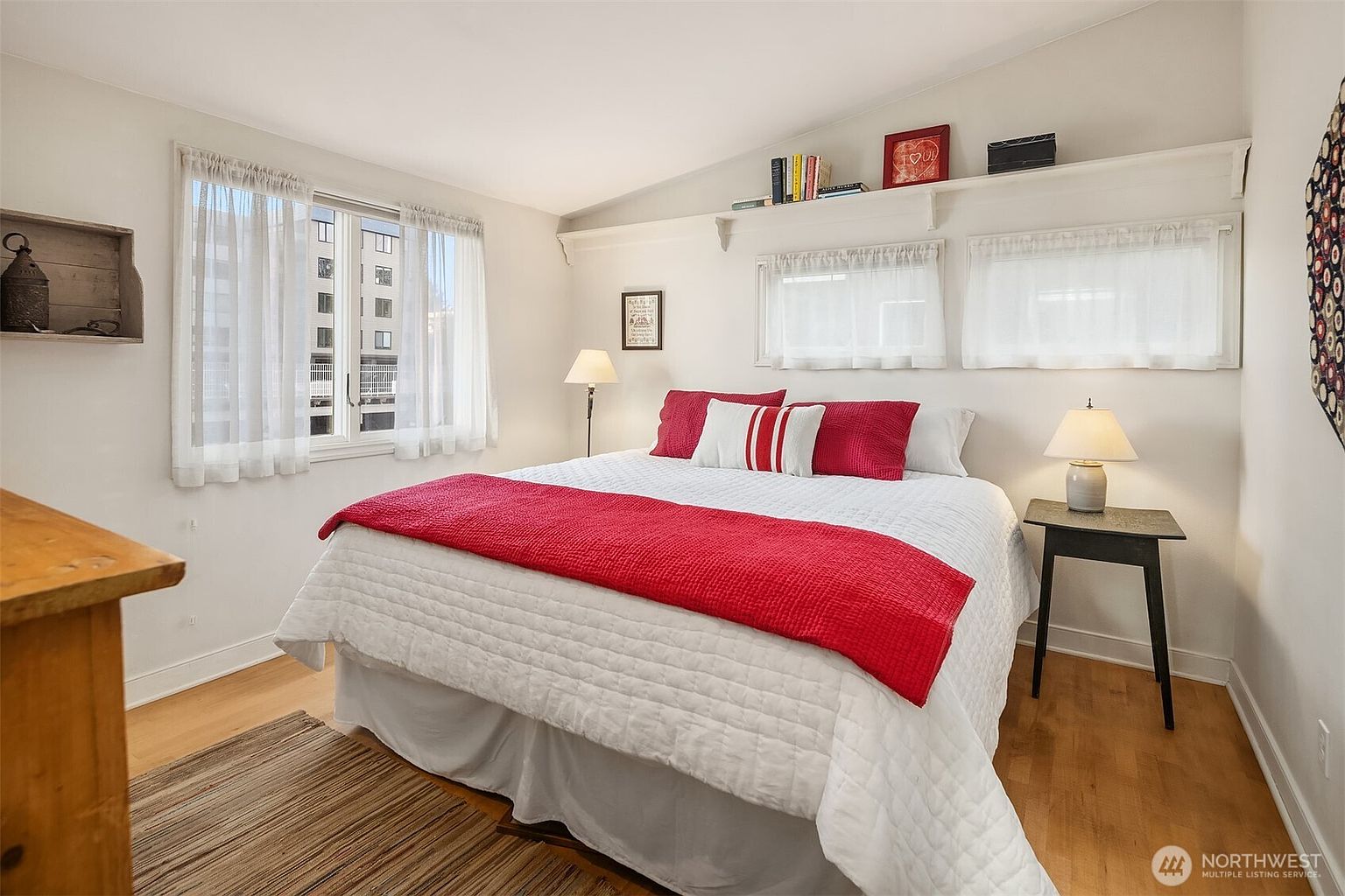 This is a bright and inviting primary bedroom featuring a large bed with white bedding and a red throw. Natural light streams in through the windows, complemented by bedside lamps. The room has hardwood floors and a minimalist style, creating a serene and comfortable atmosphere.