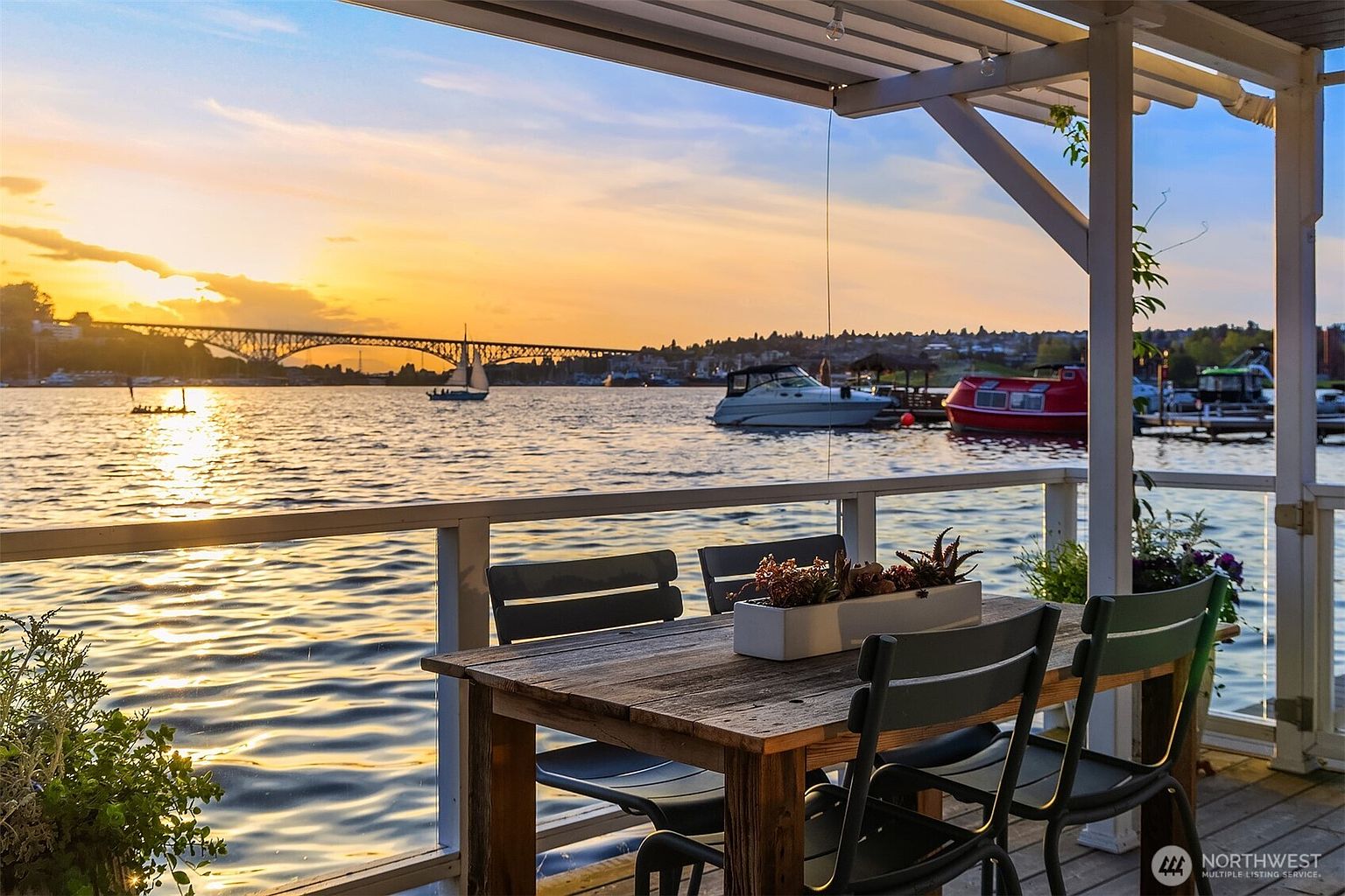 This image showcases a charming patio or deck area with a stunning waterfront view at sunset. A wooden table and chairs are arranged on the deck, creating an inviting space for outdoor dining or relaxation. The backdrop features a bridge, boats on the water, and a colorful sky, enhancing the property's appeal and highlighting its desirable location.