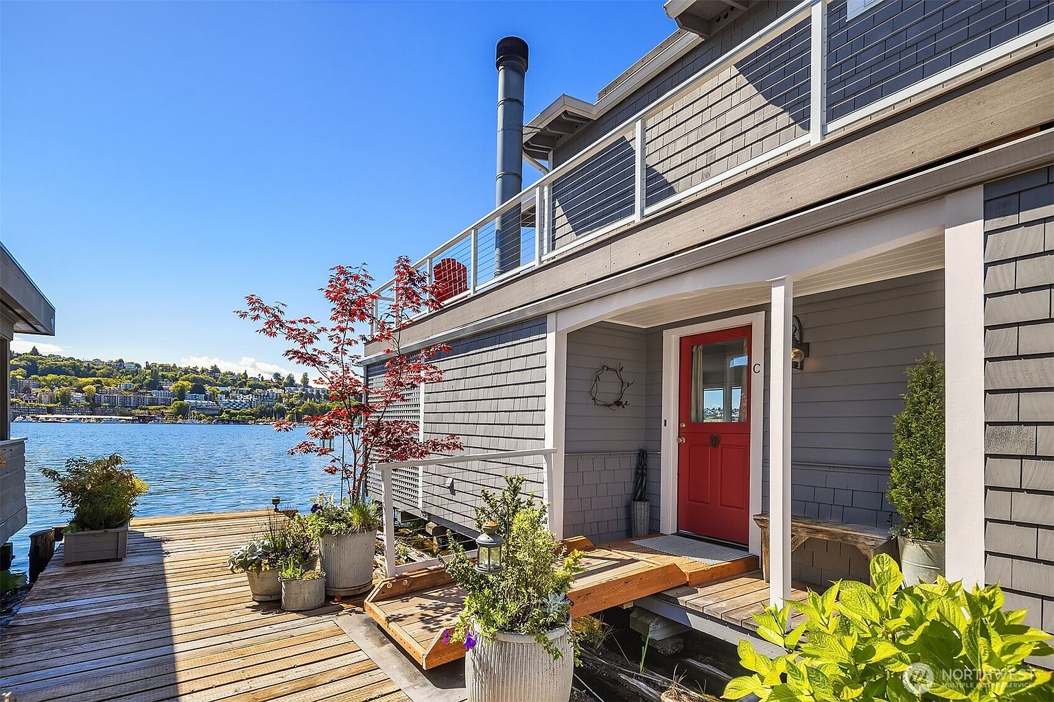 This image showcases a charming waterfront property with a wooden deck and a red front door. The house features gray shingle siding and a second-story balcony with modern railings. Potted plants add a touch of nature to the deck, creating an inviting outdoor space with a scenic water view.
